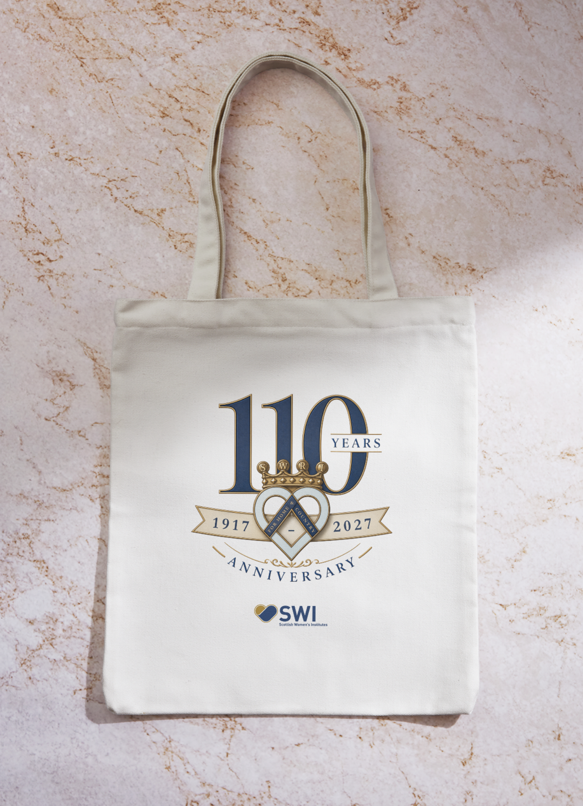A cream-colored canvas tote bag with a printed 110th Anniversary graphic for Scottish Womens Institute, displayed flat on a textured beige marble surface.